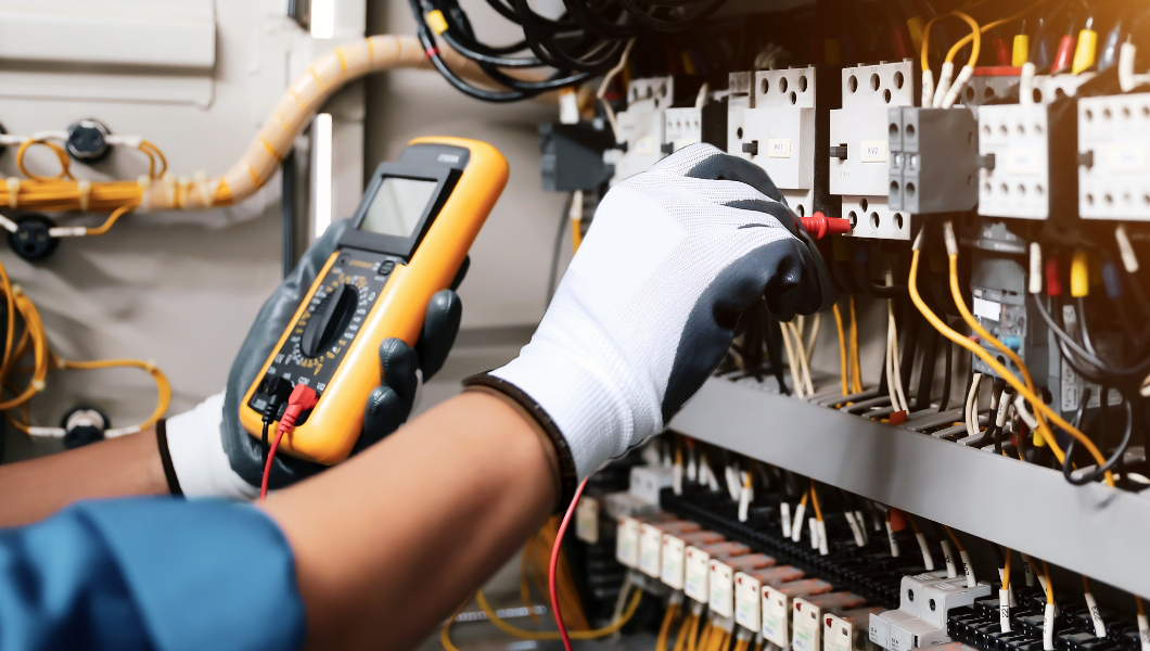 A person wearing gloves uses a digital multimeter to test electrical components inside an open control panel with various wires and switches.
