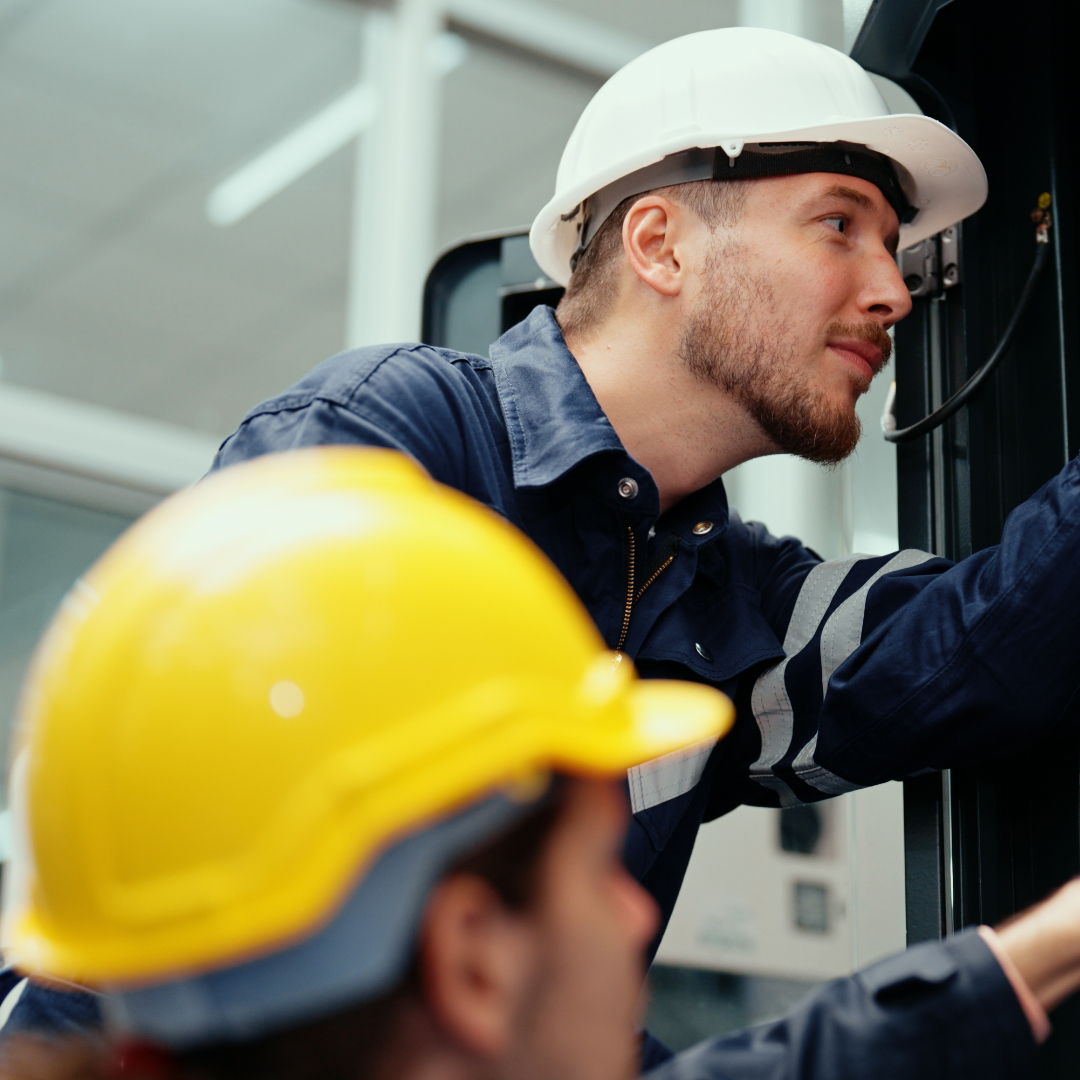 Two workers wearing hard hats, one in white and one in yellow, inspect or repair equipment, demonstrating the hands-on skills required for electrician jobs.