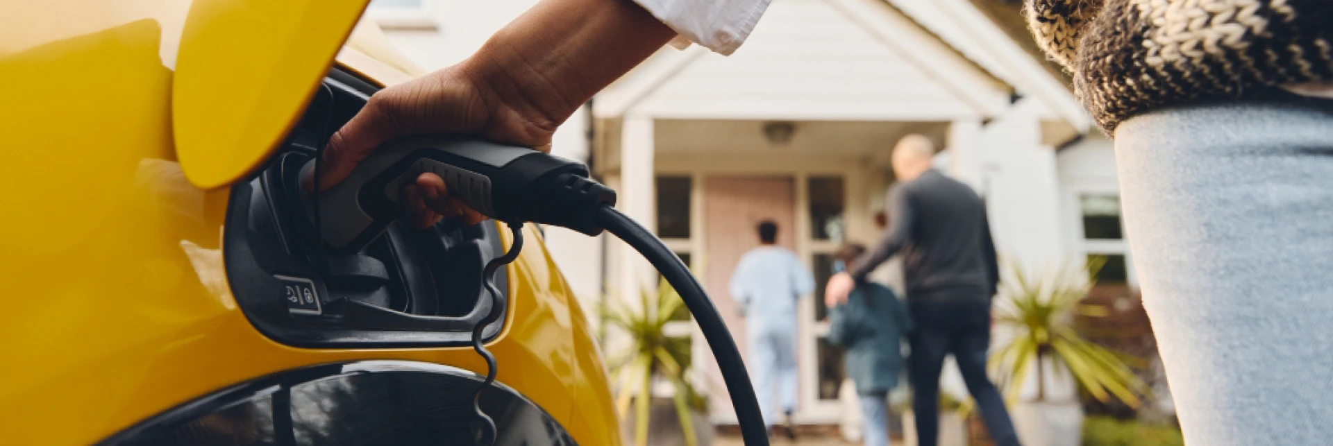 Person charging a yellow electric vehicle in a driveway, with a house and people visible in the background.
