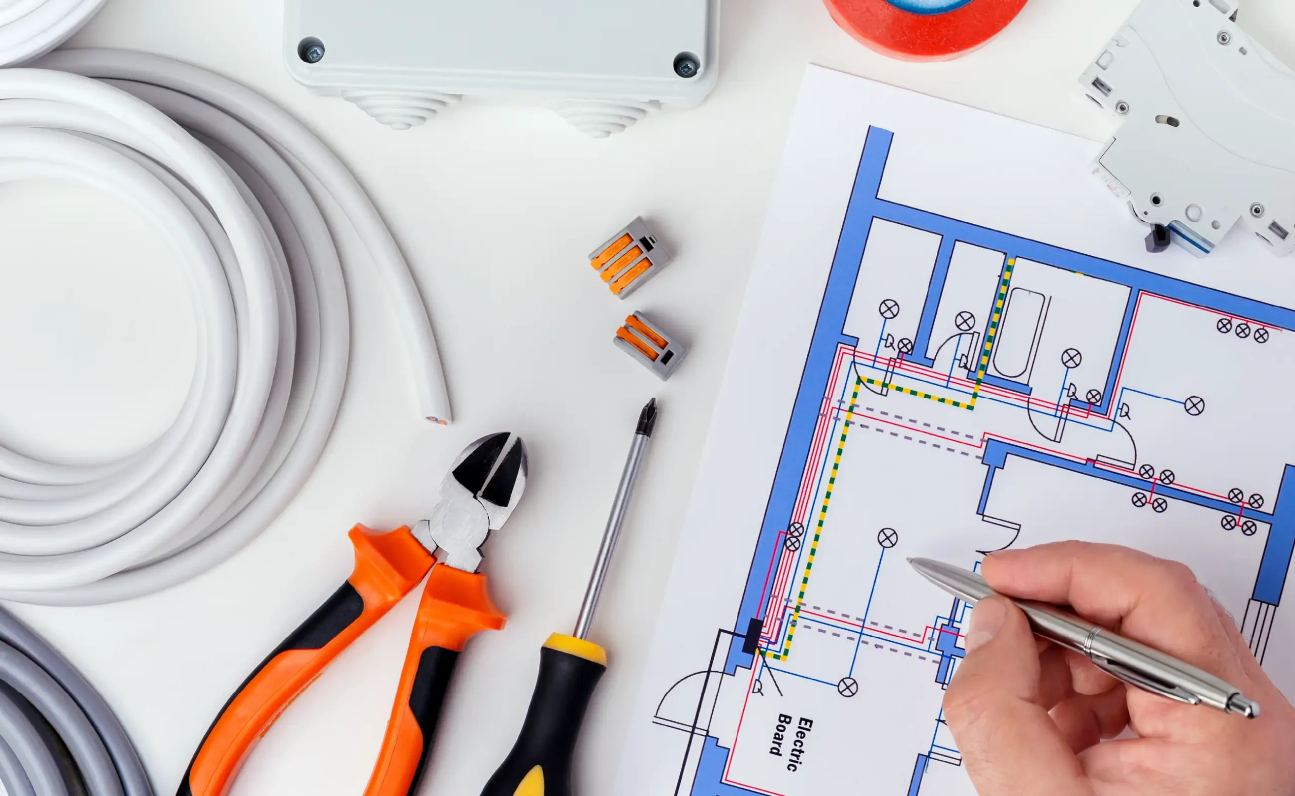 A person reviews an electrical wiring diagram with tools, wire, connectors, and tape arranged on a white surface, preparing for a home remodel.